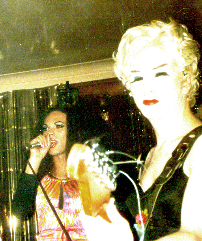 Two transfemmes perform together in front of a glitter slash curtain. Jasmine, on the left is black with long hair and wears a shiny psychedelic dress, she sings into a microphone. Mona, on the right, is bleached out, emphasising her short blonde wig, eyelashes and lipstick. She plays a guitar.