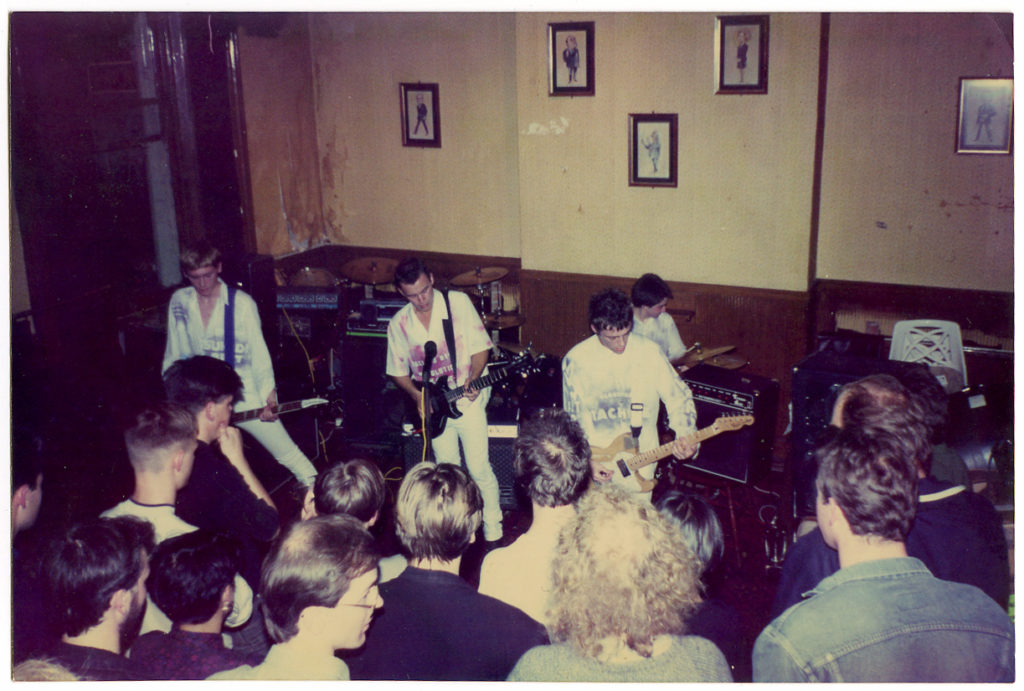 The Manic Street Preachers play a gig in a grotty pub room, they are wearing white stencilled stage outfits, backs of heads of a small crowd watching intently