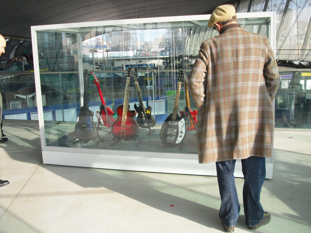 Man in checked coat and hat observes a display case of vintage guitars at Stratford Station