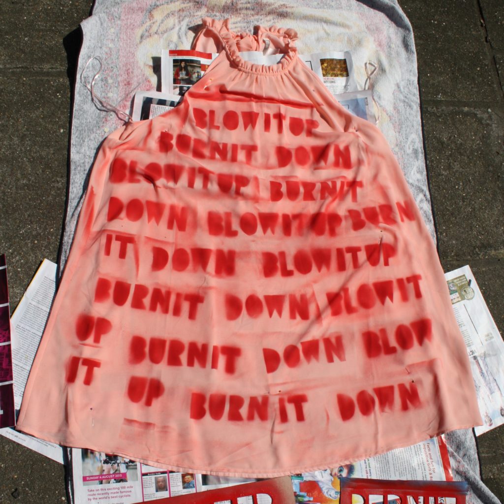 A pink polyester halterneck dress lies on a towel and paper on the floor. It has been spray-painted with a stencil that reads blow it up burn it down repeatedly
