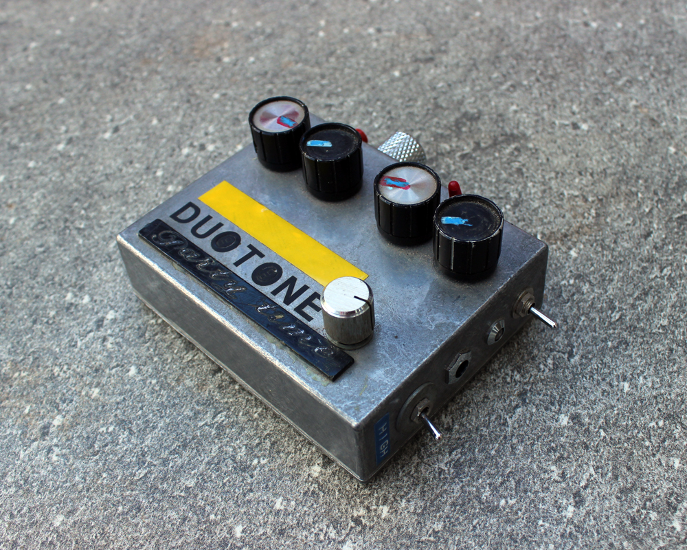 A hand made electronic pedal: small metal enclosure crowded with knobs on top, two switches on the side, inputs and outputs, a mixture of labelling with dymo tape, a plastic plaque, some yellow tape