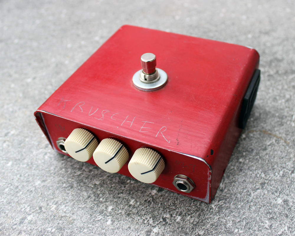 A hand made electronic pedal: red metal enclosure, one foot switch on the top, three white plastic knobs on the front, Kruscher scrtched into the metal, kind of cute!