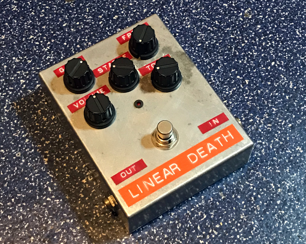 A hand made electronic pedal: boxy and metal, five knobs, a switch and dymo tape lettering