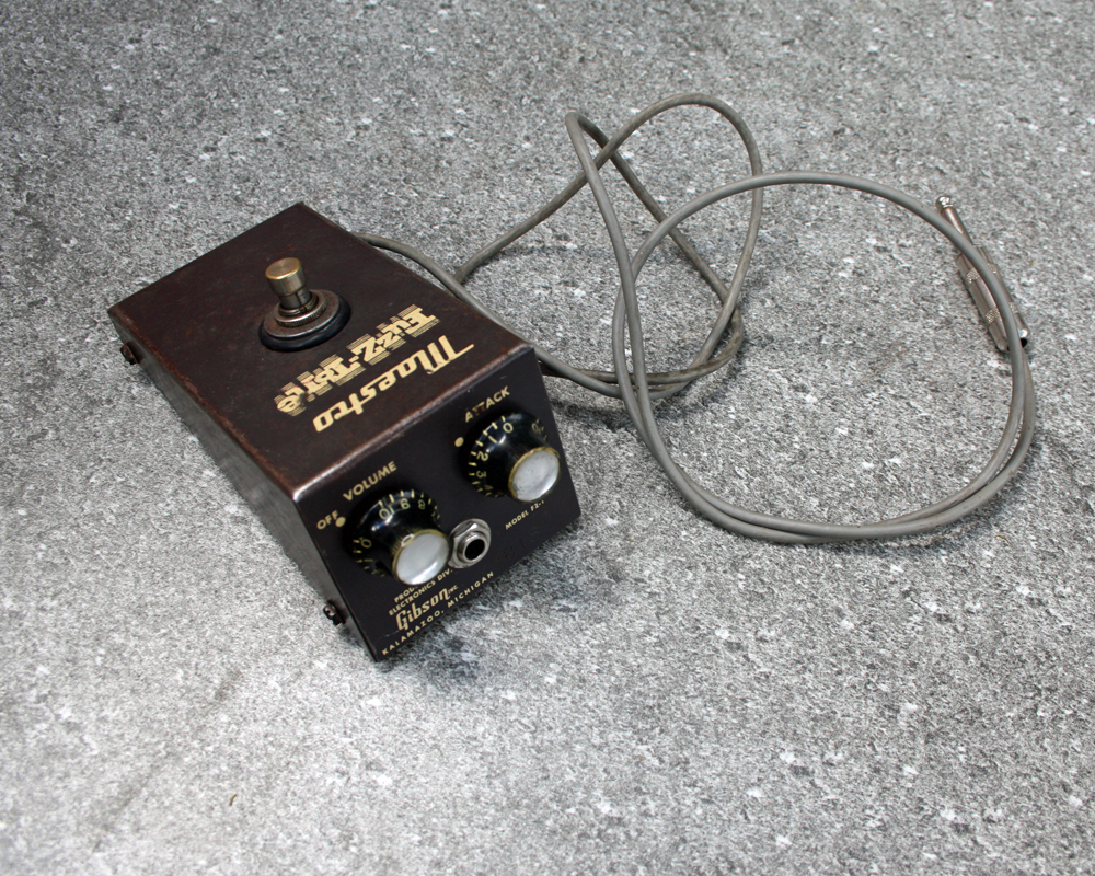 Vintage guitar fuzz pedal, rusted metal with brown paint and yellow graphics, cable attached, two knobs and an output on the front that look like a surprised face, a switch on top