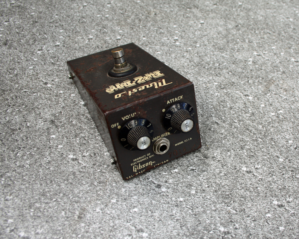 Vintage guitar fuzz pedal, rusted metal with brown paint and yellow graphics, two knobs and an output on the front that look like a surprised face, a switch on top