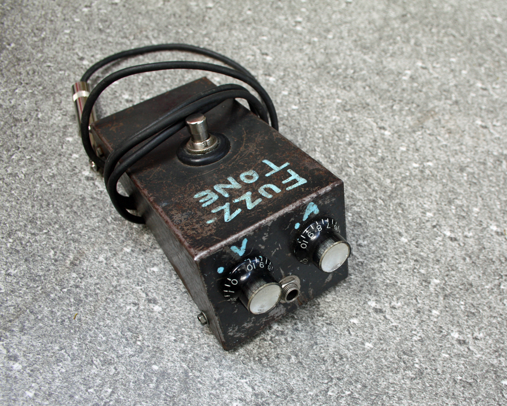 Vintage guitar fuzz pedal, rusted black metal with hand-lettered graphics, two knobs and an output on the front that look like a surprised face, a switch on top and cable attached