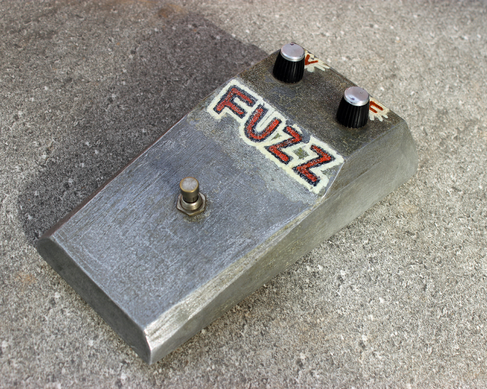 Vintage guitar fuzz pedal, metal, wedge-shaped with rough, hand-lettered FUZZ, two knobs and a switch, looks like a stone-age effects pedal