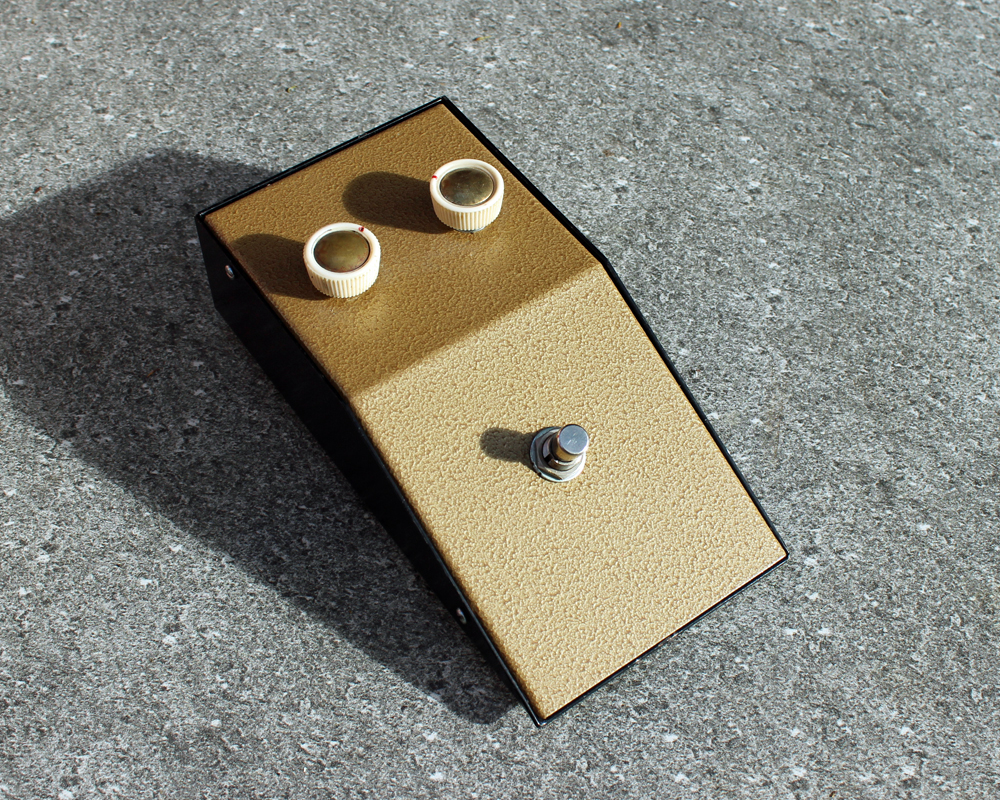 DIY guitar fuzz pedal, gold metal finish, wedge-shaped, two knobs on top and a foot switch, black metal casing, looks like a surprised robot's face