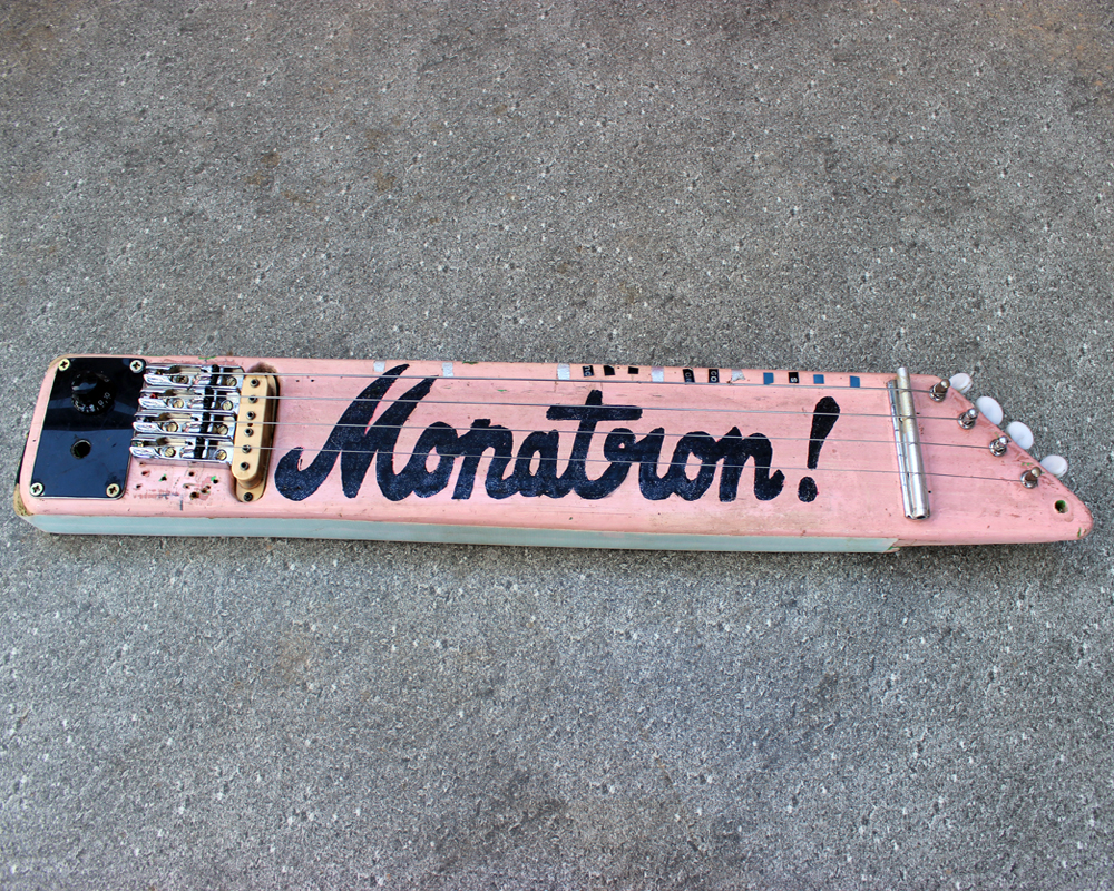 A hand-made instrument consisting of a pink plank with guitar strings on it, pick-ups, tuners. The word Monatron is painted in decorative lettering, looks well-used and fucked-up