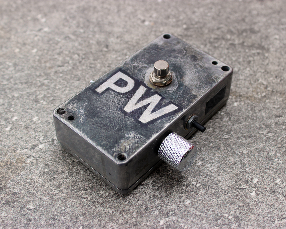 A hand made electronic pedal: metal enclosure, two switches, one knob, the letters PW drawn on it and edged in felt tip, beaten-up looking