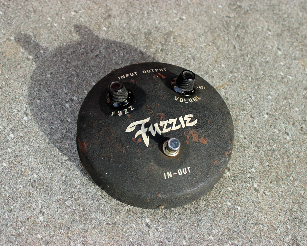 Vintage guitar fuzz pedal, round, metal, like a mic stand base, two knobs, a switch, screen-printed labels and branding, looks like a robot face
