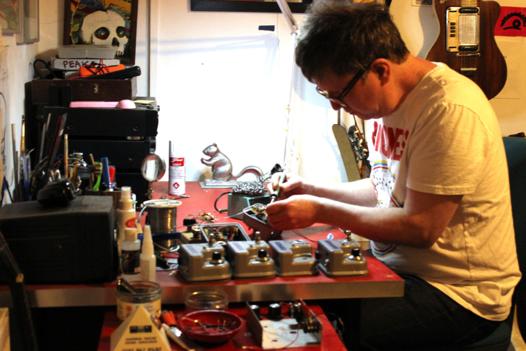 Simon Murphy making fuzz pedals at his work bench, he is surrounded by enclosures, components, tools, pictures and decorative objects. He holds a soldering iron and stares at his work intently. he has short hair, wears glasses and a Ramones t-shirt