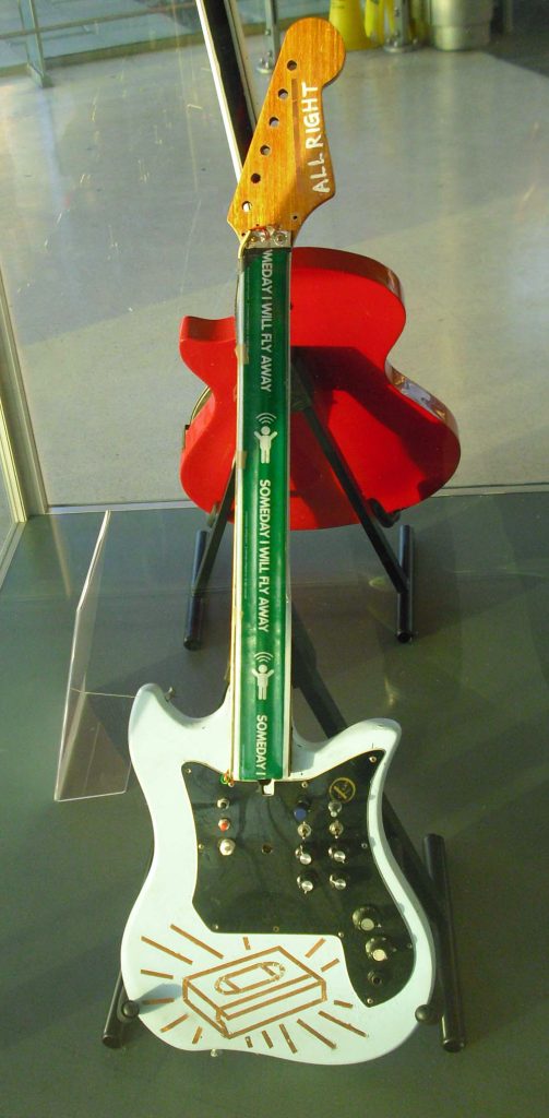 A home-made electronic instrument that looks like a guitar but has green tape along the neck instead of strings. the head has the words "All right" painted on and there is an outline of a videotape design in metal tape on the body, which is a pale blue. The plate has lots of knobs on it. The instrument is in a showcase with another red guitar behind it.