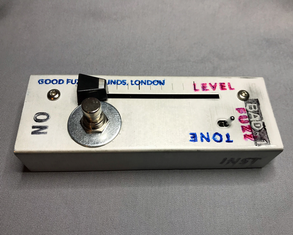 Hand-made fuzz pedal, rectangular metal enclosure with a switch and a fader. The object is printed with the words Good fuzzy Sounds, Level, Tone and Bad 1