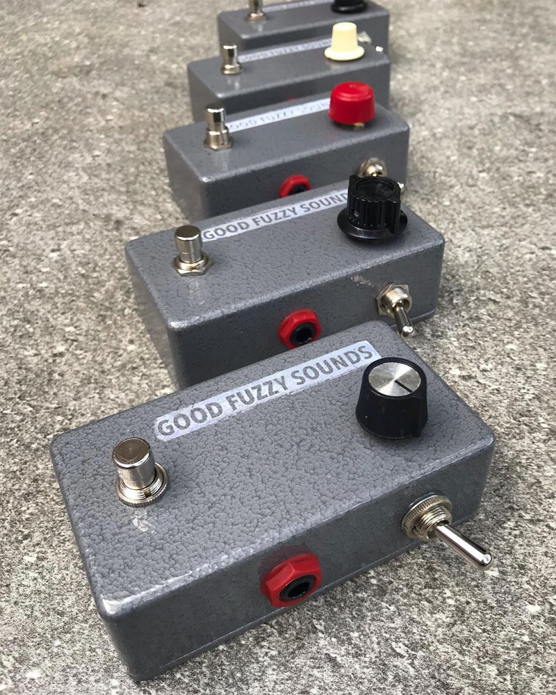 Five hand made fuzz pedals in a row, grey metal enclosures, black white and red knobs, switches, hand-stamped graphics. Pretty cute!